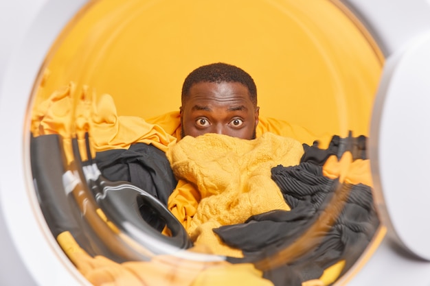  man with dark skin does laundry at laundromat hides behind pile of sorted clothes stares surprisingly poses through washing machine drum 