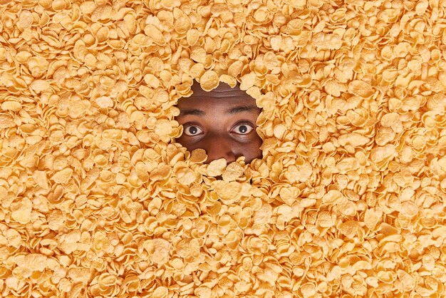  man with dark skin shows only eyes drowned in cereals has krunchy snack for breakfast makes creative shot of food ingredients. Overhead shot. Cornflakes to eat