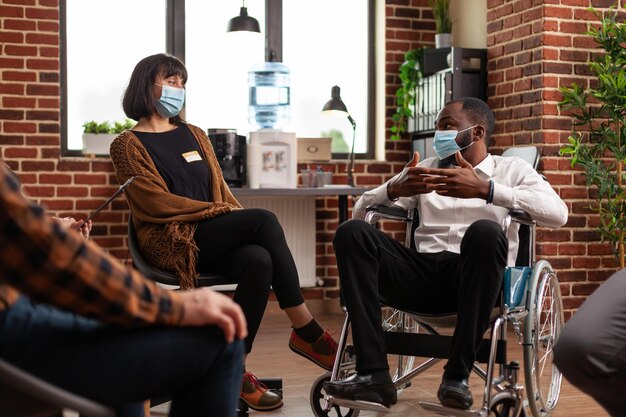 Man with disability talking at group therapy session with people having alcohol addiction. Patients wearing face masks and attending aa meeting to have discussion with psychologist.