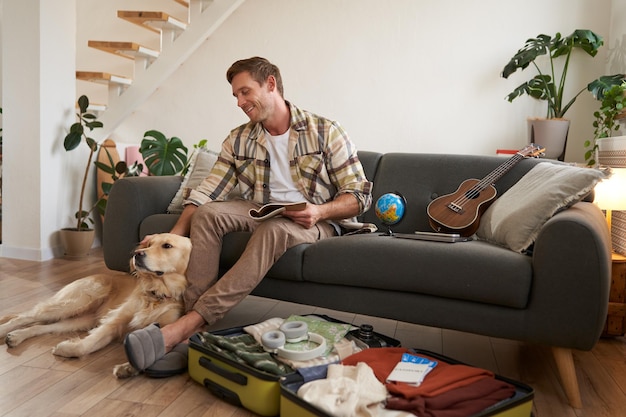 Free photo man with a dog sitting near suitcase holding notebook checking list of items to take on vacation