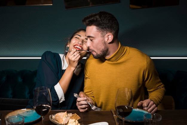 Мужчина с вилкой во рту возле жизнерадостной женщины за столом с бокалами вина и едой в ресторане