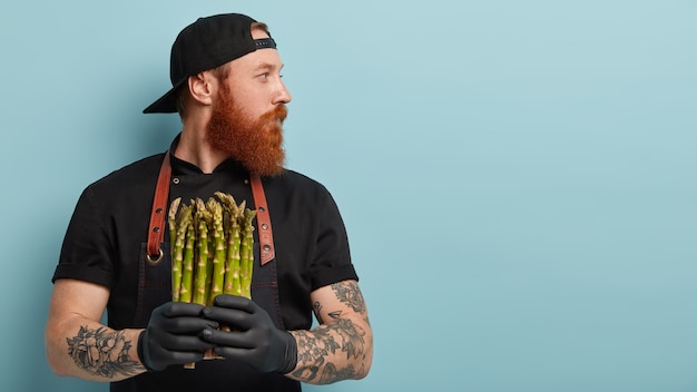 Man with ginger beard in apron and gloves holding asparagus