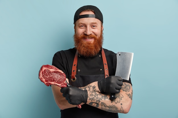 Man with ginger beard in apron and gloves holding knife and meat