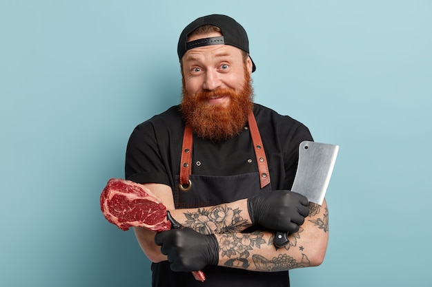 Free photo man with ginger beard in apron and gloves holding knife and meat