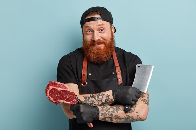 Мужчина с рыжей бородой в фартуке и перчатках держит нож и мясо