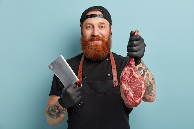 Free photo man with ginger beard in apron and gloves holding knife and meat