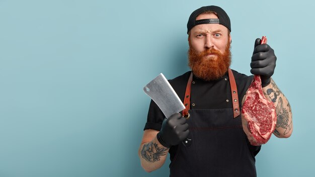 Man with ginger beard in apron and gloves holding knife and meat