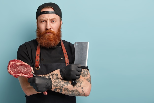 Man with ginger beard in apron and gloves holding knife and meat