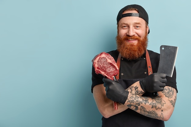 Man with ginger beard in apron and gloves holding knife and meat