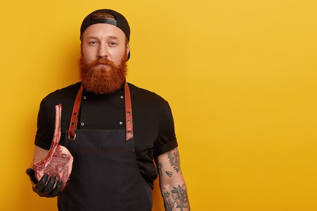 Мужчина с рыжей бородой в фартуке и перчатках держит мясо