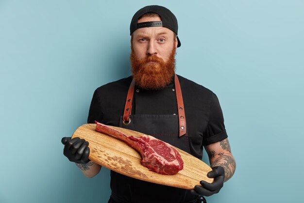 Free photo man with ginger beard in apron and gloves holding wooden board with piece of meat