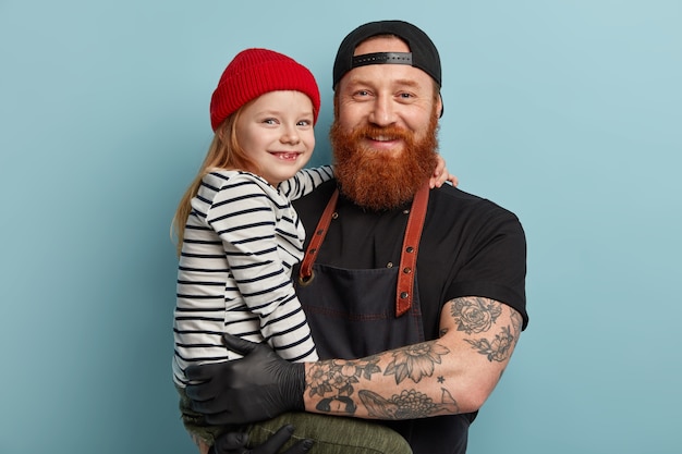 Free photo man with ginger beard wearing apron and gloves holding his daughter