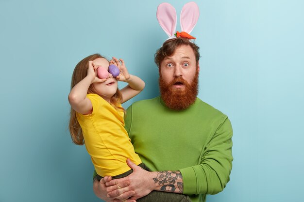 Man with ginger beard wearing colorful clothes and bunny ears holding his daughter