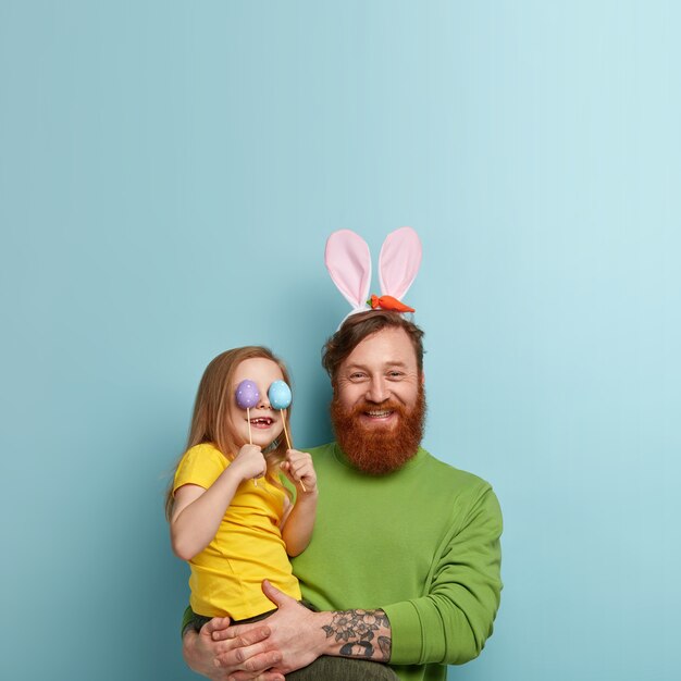 Man with ginger beard wearing colorful clothes and bunny ears holding his daughter