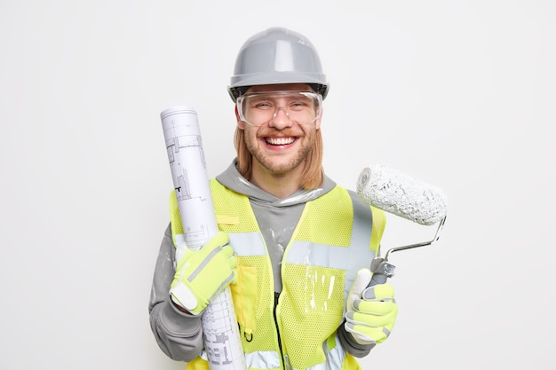 man with ginger hair holds paper blueprint and painting roller dressed in protective helmet safety traansparent glasses uniform isolated on white