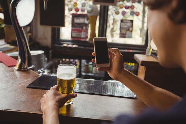 カウンターで携帯電話を使用してビールのグラスを持つ男