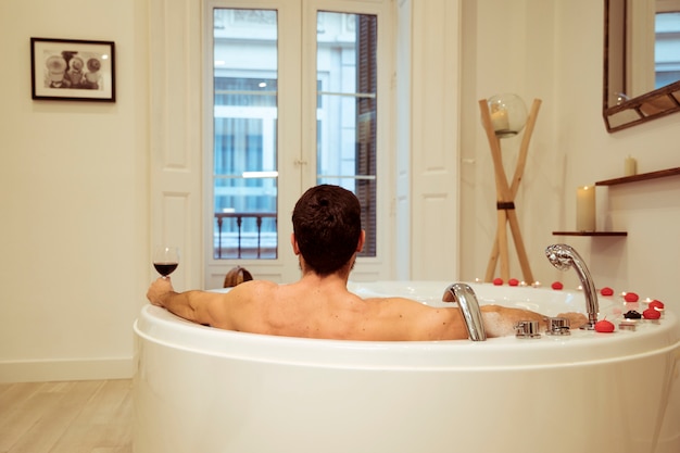 Man with glass of drink in spa tub with burning candles