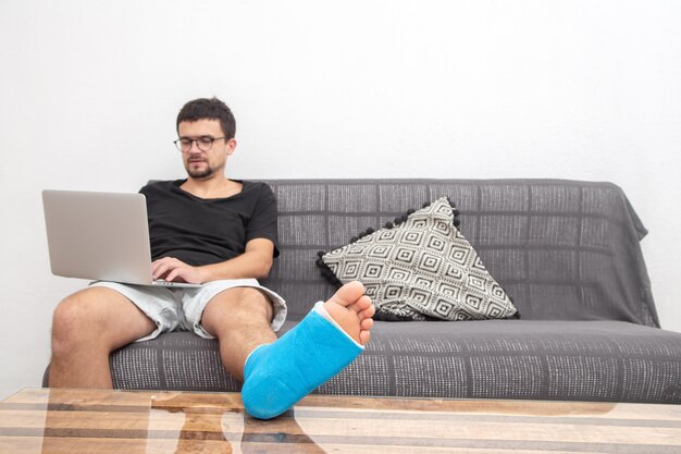 Man with glasses with broken leg in blue splint for treatment of injuries from ankle sprain working on a laptop on couch at home.