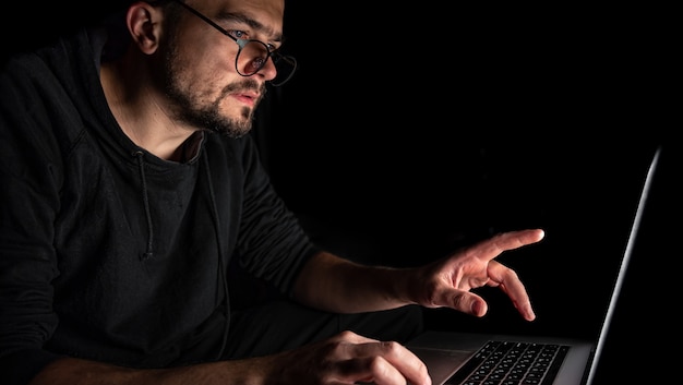 A man with glasses works at a laptop in the dark