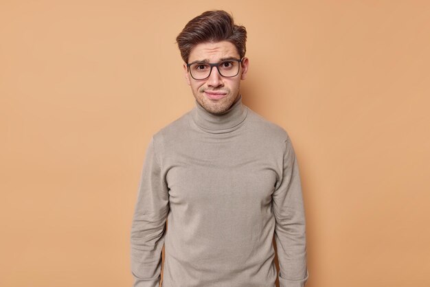 man with hairstyle looks unhappily at camera reacts on bad news wears transparent eyeglasses and casual poloneck isolated on beige. Negative face expressions.