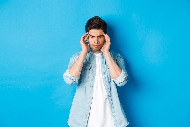 Man with hangover touching head and grimacing, having headache, standing against blue background