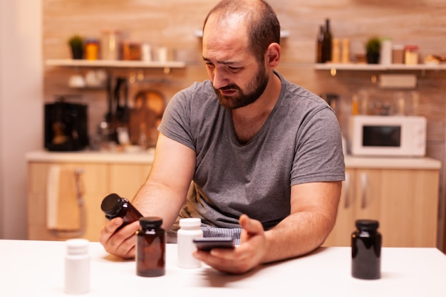 Man with headache holding pills bottle sitting in the kitchen. Stressed tired unhappy worried person suffering of migraine, depression, disease and anxiety feeling exhausted with dizziness symptoms.