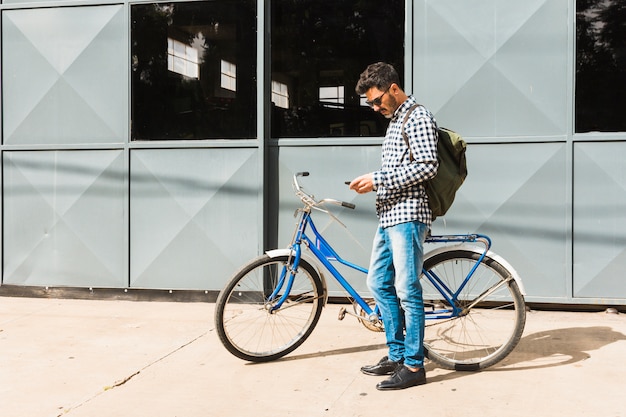 自転車の近くに立っている携帯電話を使用して彼のバックパックを持つ男