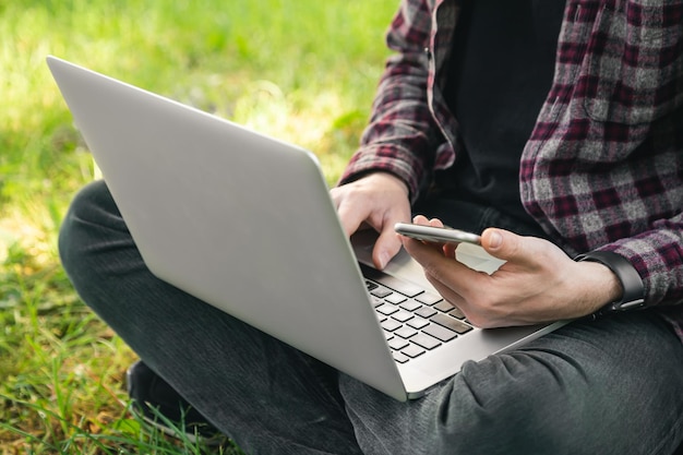 Foto gratuita un uomo con un computer portatile e uno smartphone si siede sul primo piano dell'erba