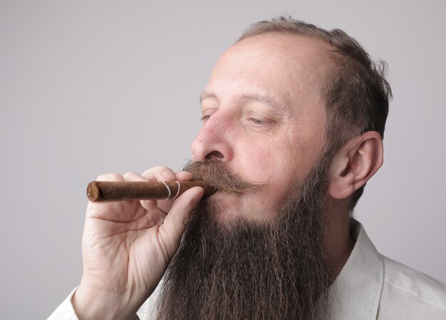 Человек с длинной бородой и усами курит сигару с серой стеной