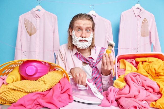 man with long red hair busy doing ironing of clothes and shaving at same time being in hurry as onslept work