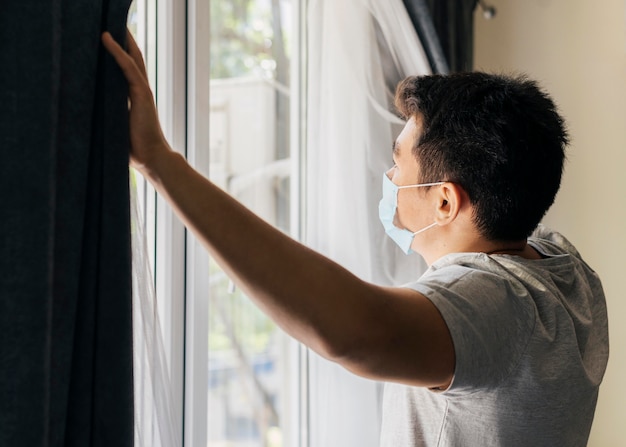 Человек с медицинской маской дома во время пандемии открывает оконные шторы