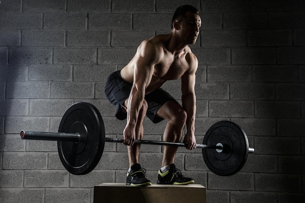 Man with naked torso holds barbell over the grey wall