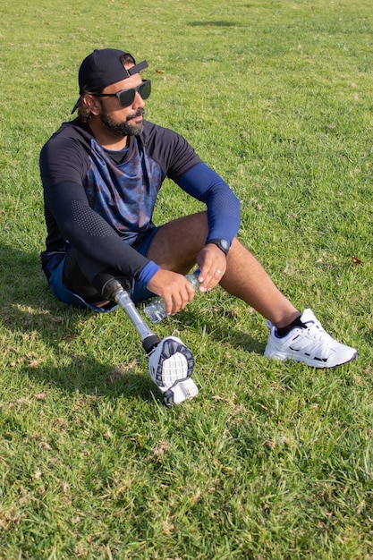 Man with prosthetic leg relaxing after training. Caucasian man with beard drinking water after exercises, sitting on grass. Sport, leisure, disability concept
