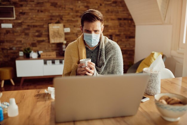 Man with protective face mask having a cup of tea while using laptop at home