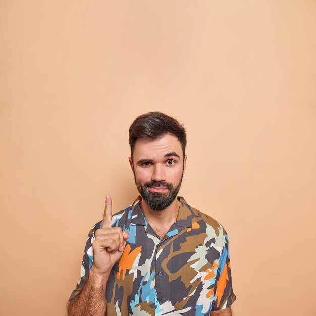 L'uomo con l'espressione sicura di sé indica verso l'alto mostra il banner promozionale consiglia di andare di sopra indossa una camicia colorata posa su beige