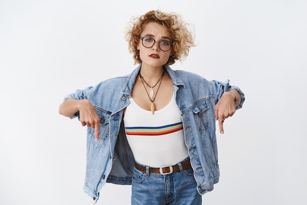 Free photo man with short blond hair in transparent glasses and denim jacker pointing down with index fingers and looking unsure, hesitant as asking question.