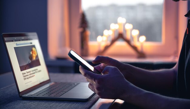 A man with a smartphone sits in front of a laptop late at night