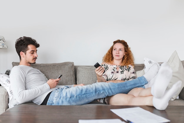 Человек со смартфоном, сидя рядом со своей женой, смотрит телевизор