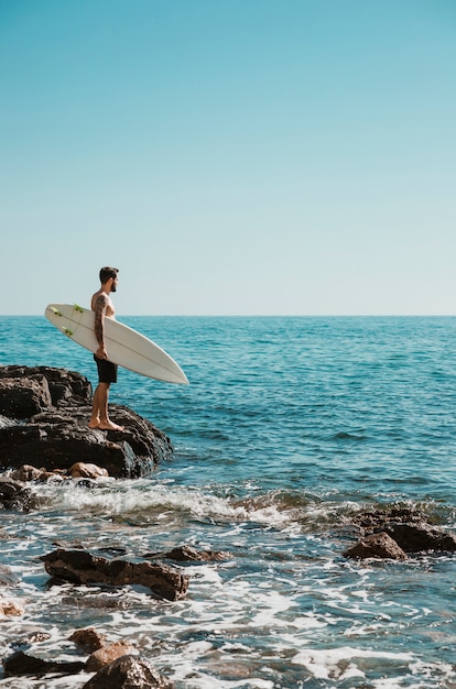 石の多い海岸に立っているサーフボードを持つ男