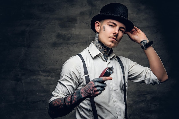 A man with tattoo on a face, dressed in a white shirt and a cylinder hat holds hair trimmer.