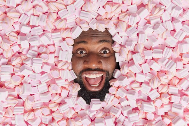 man with thick beard looks happily keeps mouth opened drowned in sweet appetizing marshmallow enjoys eating tasty yumminess