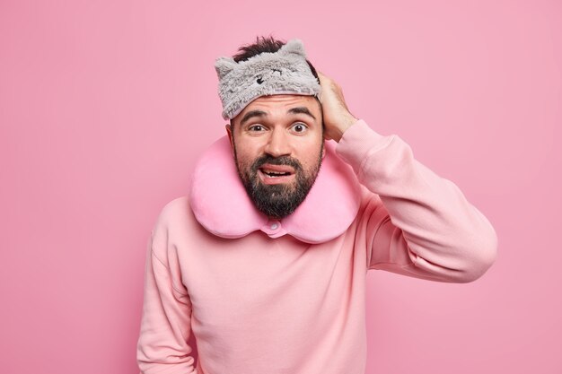  man with thick beard scratches head frowns face wears sleepmask neck pillow dressed in casual jumper 