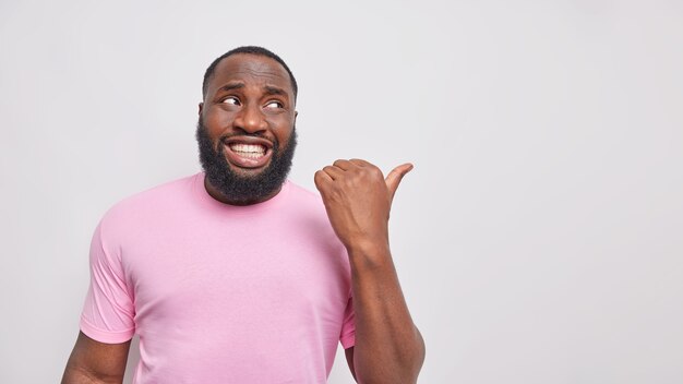  man with white teeth points thumb away on blank space shows place for logo or product placement dressed in casual pink t shirt 