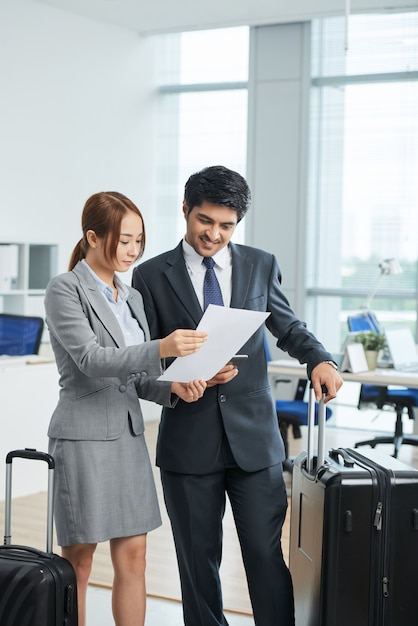 Man and woman in business suits standing in office with suitcases and looking together at document