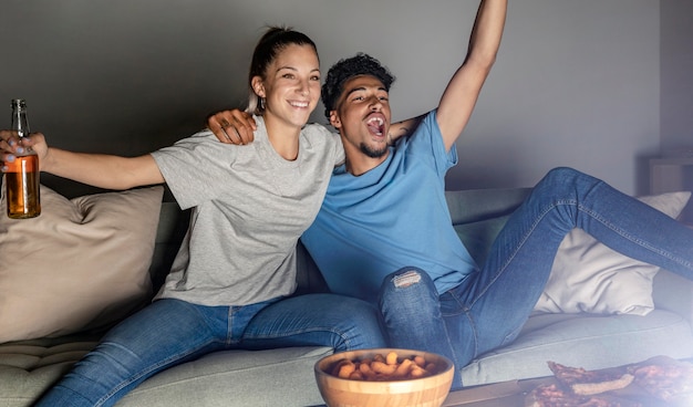 Man and woman having beer and snacks at home while watching tv and cheering