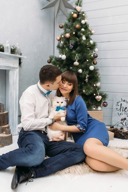Man and woman hug each other posing with white kitty before a Christmas tree