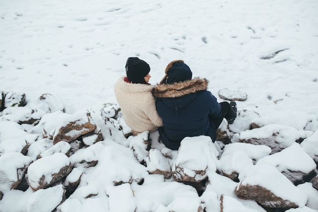 Man and woman sitting on the rocks covered with snow, on the shore of the lake