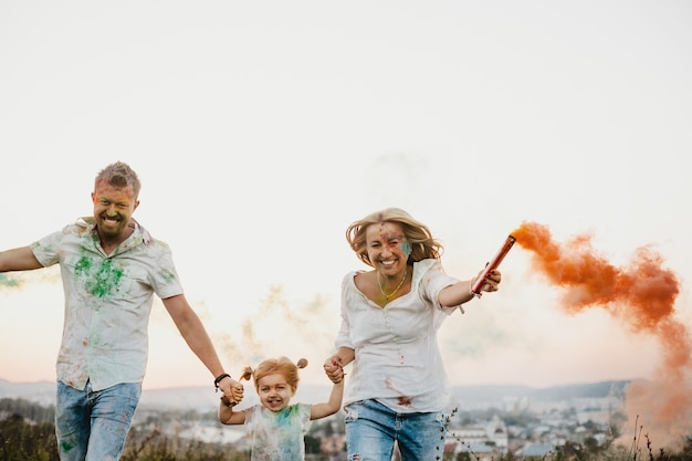 Foto gratuita l'uomo, la donna e la loro piccola figlia si divertono a correre con il fumo colorato tra le loro braccia