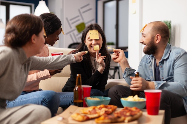 Man and women enjoying guessing game with sticky notes, playing charades together at drinks celebration after work hours. Office workmates having fun with patnomime activity at party.