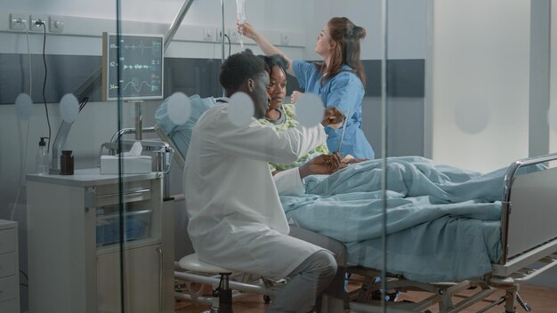 Man working as medic and explaining x ray scan results to patient with IV drip bag in hospital ward bed. Medical specialist holding radiography to talk to young woman about healthcare